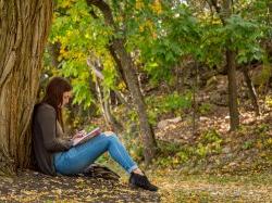 Photo of a student leaning against a tree and writing in a notebook